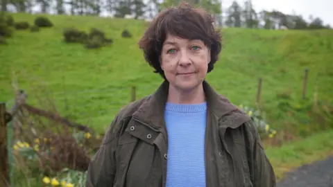 Natalie Barstow, lead claimant, stands on her farm near Builth Wells in Powys.  She has brown hair, blue eyes and is wearing a light blue jumper and dark green coat.  Green fields and daffodils can be seen behind her.