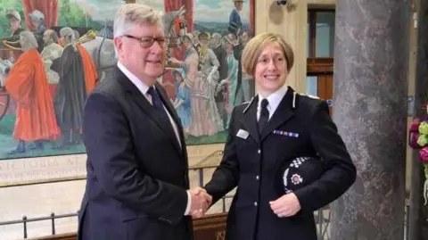 ESSEX PFCC Rachel Nolan, on the right, shaking hands with Roger Hirst, the Police, Fire and Crime Commissioner for Essex. They are standing next to a marble pillar inside County Hall in Chelmsford. There is a large wall painting showing a crowd scene behind them. Hirst is in a dark suit, a white shirt and a blue tie. Nolan is in her police uniform and has her hat under her left arm.