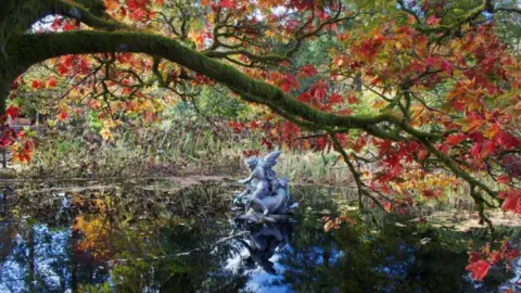 Caitlin McGuire Un estanque sereno que refleja el follaje otoñal, con una rama de árbol cubierta de musgo arqueada en lo alto y una estatua de una figura alada en el agua.