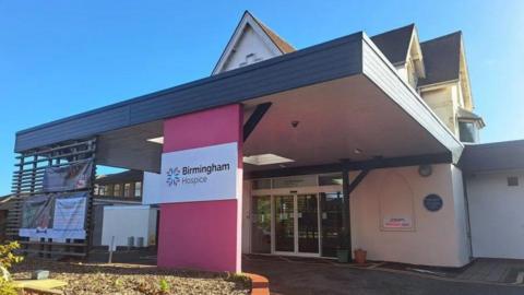 The entrance to Birmingham Hospice with a bright pink pillar which supports a canopied area with glass doors and a brick building behind