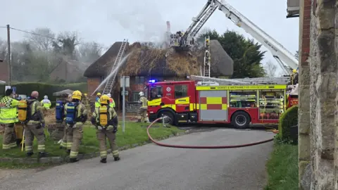 A fire engine with red and yellow hi-vis decals parked in the middle of the road. There are around nine firefighters standing nearby - one spraying water from a hose onto the thatched roof cottage, which is visible in the distance. Smoke can be seen coming from the destroyed roof, which appears burnt and pulled apart.