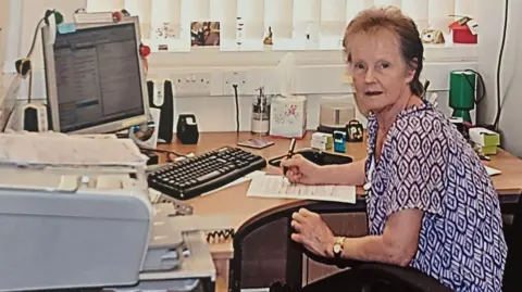 Yvonne Clark looks directly at the camera as works on paperwork at a desk in a conservatory. She is wearing a blue and white top and has a gold watch on her left arm. Her desk is filled with office supplies, including a computer, printer and keyboard. She is sitting sideways to the camera on an office chair.