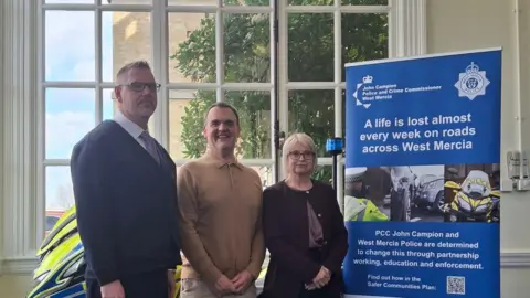 West Mercia Police and Crime Commissioner The photo shows three people standing indoors in front of a large arched window. Behind them, greenery can be seen outside through the glass.
To the left of the group, part of a high‑visibility police motorcycle is visible, including bright yellow and blue markings.
To the right of the group, there is a tall, freestanding display banner. The banner features the West Mercia Police logo and contains road‑safety messaging. The main headline on the banner reads:
“A life is lost almost every week on roads across West Mercia.”