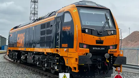 The new locomotive is on a set of tracks. It is very clean and shiny with an orange front and black sides. On the side, written in orange is 'GBRF'.