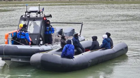 Two boats are seen in a canal - in the foreground is a grey inflatable with six people sitting inside. In the background is a police boat with men in light blue jackets.