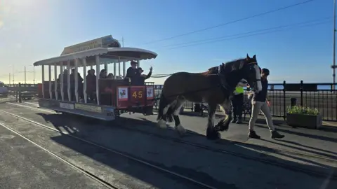 BBC A brown horse pulls a Victorian tram car full of passengers, with railings along the road behind and unbroken blue skies.