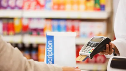 Getty Images A chemist handing a card machine over to a customer who is buying a prescription. The gold card is in the silver machine and the prescription is in a white bag with a blue stripe. The background of medicines is blurred.