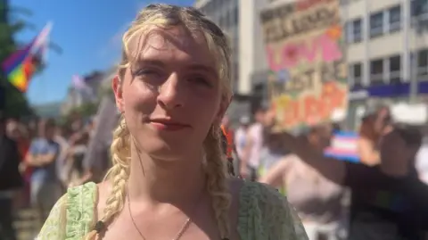 Alexa is smiling at the camera. She has long blonde hair which is in two plaits. She is wearing a green top.