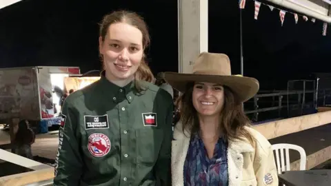 Woolley family Two women stood in the sheltered outdoor area of a bar. Left, the woman is wearing a green shirt with embroidered badges on. Right, the woman is wearing a large brown cowboy hat, cream coat and blue blouse.