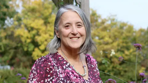 Farleigh Hospice A head and shoulders image of Michelle Kabia. She is wearing a pink, burgundy and white speckled top and a necklace. She is standing in front of green bushes and trees with purple flowers. She is smiling and looking into the camera.
