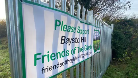 The metal fencing, with spikes on the top, is stretching diagonally across the image. On the fencing, a banner, reading 'Please Support Bayston Hill Friends of the Glebe' has been placed - it's also got their email address, friendsoftheglebe49@gmail.com, written on it. The lettering is green and black on a white background. You can see a bit of the field behind the fence - it's early morning so the light is pink and slightly sunny. 