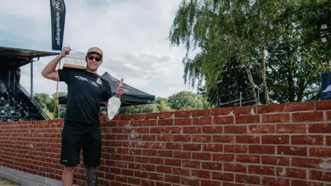 A man in a black tshirt and cap is standing next to a brick wall holding a trowl. 