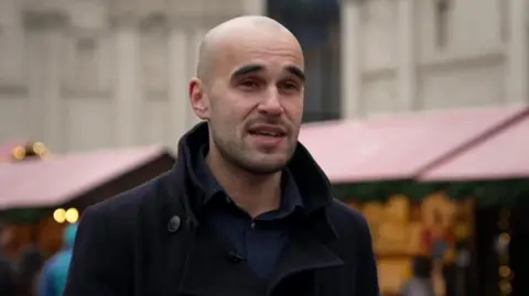Simeon Scheuber-Rush is pictured in Trafalgar Square, with wooden Christmas market stalls visible behind him.