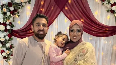 Akeel Nanabawa and Hannaa Vorajee hold their daughter Sara at a celebration event. Akeel wears a grey top while Hanaa wears a terracota head scarf and gold top. Sara is dressed in pink and they stand in front of a floral display