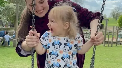 Instagram Isabella Jonas-Wheildon, a blonde-haired toddler, smiles while sitting on a swing. Her mum, Chelsea Gleason-Mitchell - a woman with brown hair and a burgundy top is smiling behind her. The child is wearing a top with Minnie Mouse's face on it.
