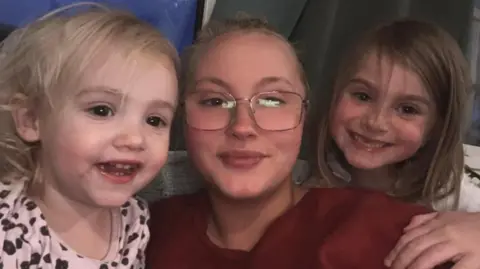 Natasha Major Natasha is smiling and wearing a read top and has her two daughters next to her, one on either side of her, also smiling