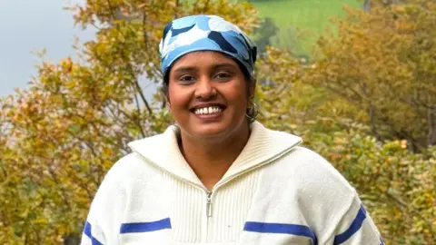 Rafiya Sherin Woman wearing a blue and white bandana and a white wool pull-over. She is smiling standing outside on a hillside.