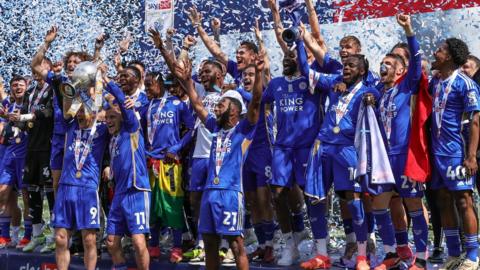 The Leicester squad lift the Championship trophy together as tape falls down behind them