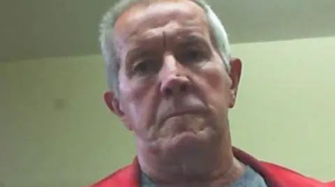 PA Media Richard looks down at a camera in a grainy picture against a green background. He has a stern expression and short grey hair, and wears a bright red jacket. 