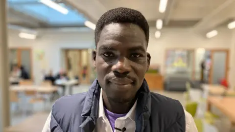 BBC Refugee Abdullah Zaeed from Sudan, who has short-cropped hair and is wearing a black gillet and white shirt, stood in hall. 