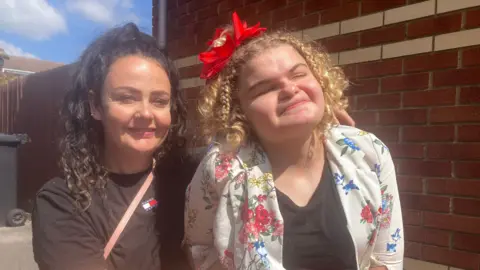 Two women pictured next to each other. The woman on the right, with light coloured hair has her eyes closed and a red decoration in her hair. The woman on the left has darker hair and is wearing a dark shirt.