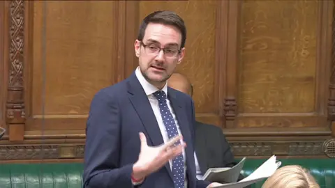 House of Commons John Aldridge speaking at the Houses of Commons. He has dark brown hair, glasses and wears a white shirt and navy blue suit.