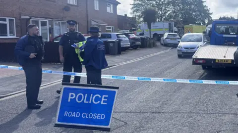 A police cordon and 'road  closed' sign on a residential street. Two PCSOs are talking to a police officer who is standing on the other side of the cordon.