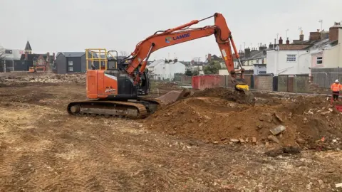 An orange digging machine on a brownfield site, where some rows of houses can be seen in the background.