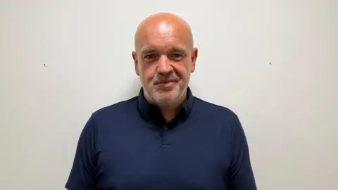 Sam Burton/BBC A man wearing a dark blue polo shirt smiles at the camera in front of a plain white wall