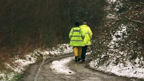 PA media dua petugas polisi dengan jaket hi-vis berjalan menyusuri trek negara. Permukaan ditutupi dalam campuran lumpur dan diaduk salju. Ada semak -semak di kedua sisi jalan.