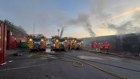 Three fire engines and firefighters at Tulketh Industrial Estate tackling a fire at commercial units. Smoke can be seen rising in the air.
