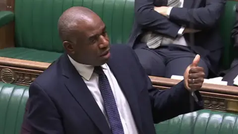 UK Parliament David Lammy speaking in the House of Commons. He is wearing a navy blue suit and tie and holding his thumb up.