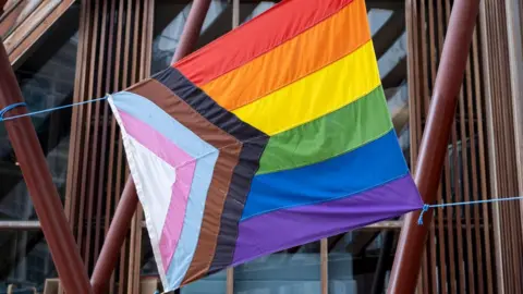 Getty Images A pride progress flag hanging from a building. It is suspended by blue string tied to its corners. It is made up of five colours in an arrow on the right, which intersect with six horizontal stripes made up of different colours of the rainbow.