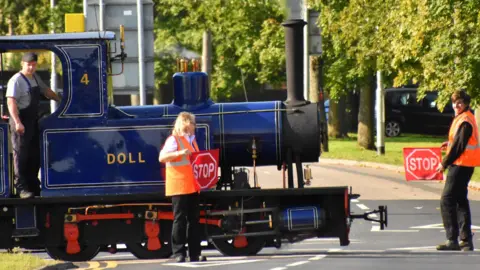 Two volunteers, standing in a road, wearing orange high-vis jackets and holding two stop signs. A man in a cabin of a blue steam train is by them. The train is crossing a road, with trees behind them. 