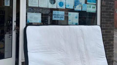 Sea Adora A large mattress fly-tipped outside of the charity shop. The mattress is white and there are signs/leaflets in the window. One of the signs says 'volunteer with us'. There is a door to the left.