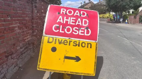 BBC A bright red "road ahead closed" sign, on top of a bright yellow "diversion" sign on a road. There is a red brick wall on the left hand side of the signs, and there are a few roofs visible in the background.