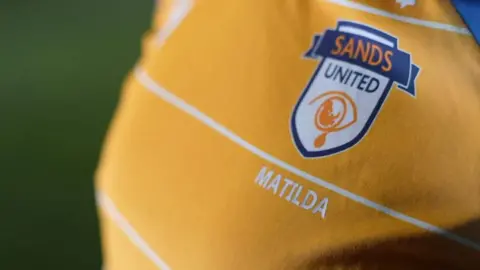 Jamie Niblock/BBC A yellow football jersey with white stripes and a Sands United crest. Under the crest is the name Matilda.