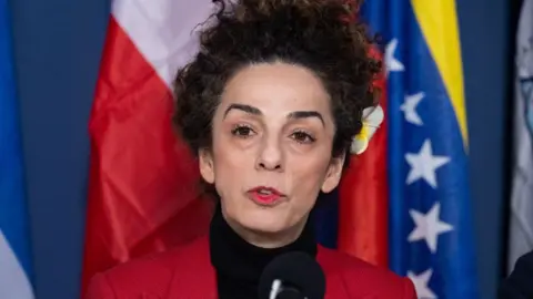 Masih Alinejad stands in front of several flags with a black shirt and red jacket. She has a flower in her hair. 