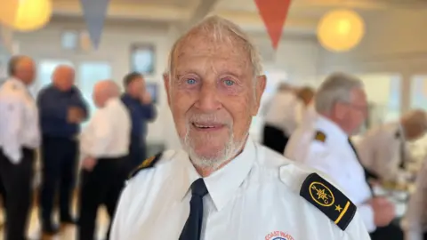 George Carden/BBC An elderly man with short white hair is smiling at the camera. He is wearing a white shirt with black lapels on the shoulder, and a black tie