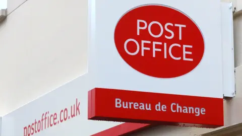 A white building with a white and red sign, pointing out the Post Office. The sign says POST OFFICE in white writing within a red oval and beneath that it reads, Bureau de Change.