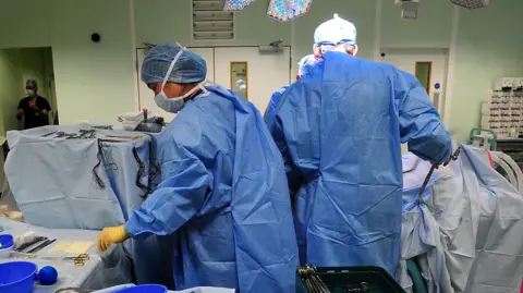 PA A female surgeon in a blue theatre gown is turning to pick up a surgical instrument during an operation. Another surgeon is standing next to her