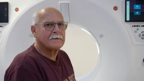 Jamie Niblock/BBC David Catchpole with short hair and glasses. He is wearing a dark T-shirt. He is sitting in front of a CT scanner.