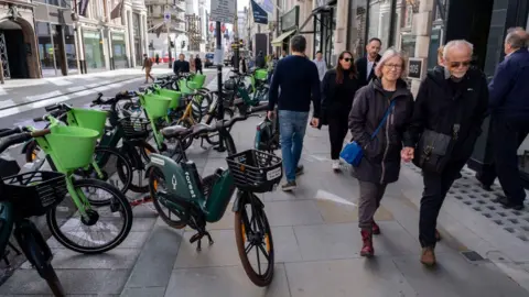 Getty Images Lime and Human Forest e-bikes parked across the pavement along Bond Street, London. People walk past avoiding the bikes.