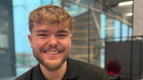 Saffron Humphreys/BBC A head and shoulders images of a man with blond short hair and a beard. He is smiling and wearing a black polo shirt.