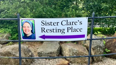 A sign reading: ' Sister Clare's Resting Place' and a purple arrow pointing left is attached to a black railing. A number of trees and stones can be seen in the background.