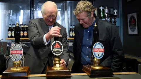 Reuters King Charles, wearing a grey suit, stands behind a bar and pulls a pint while a man wearing a black jacket and blue shirt stands next to him and watches.