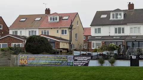A row of semi-detached houses with a stretch of grass in front. One of the houses has a banner on it's fence which reads 'Say No to South Tyneside Council putting forty lamp-posts along this route'.