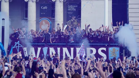 PA Media Villa players on a bus lift the trophy as fans raise their arms below.