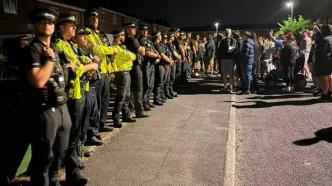 People are gathered in the street outside a property. A row of police officers are visible outside the house. Some of the people are wearing hoodies and holding mobile phones.