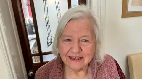 John Devine/BBC Marilyn Batrick has short grey hair swept across her head. She is wearing a bobbly, woolly light pink fleece top, behind her is a front door and some pictures hanging on the wall.